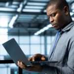 A man stands in a large, open data center. He is holding an open laptop and looking intently at the screen.