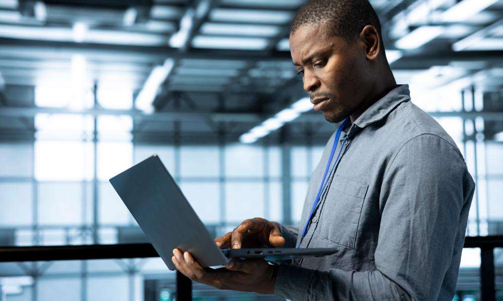 A man stands in a large, open data center. He is holding an open laptop and looking intently at the screen.