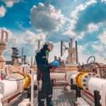 A male worker wearing protective gear inspects a long steel pipe and a pipe elbow at an oil station factory.