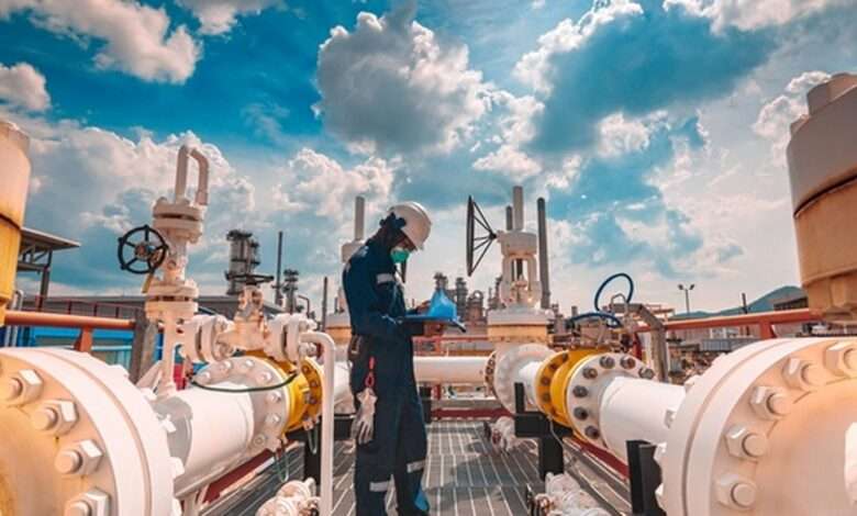 A male worker wearing protective gear inspects a long steel pipe and a pipe elbow at an oil station factory.