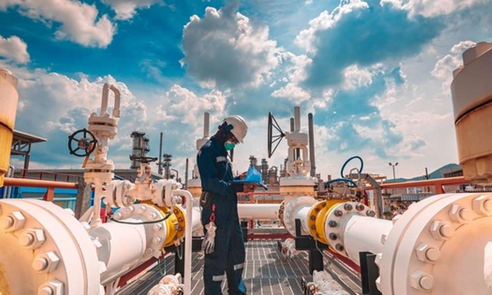 A male worker wearing protective gear inspects a long steel pipe and a pipe elbow at an oil station factory.
