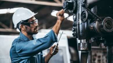 A man wearing a white hard hat, safety goggles, and blue shirt pulls down the lever on an industrial equipment.