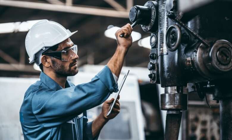 A man wearing a white hard hat, safety goggles, and blue shirt pulls down the lever on an industrial equipment.