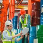 Two workers in hard hats and safety vests stand next to liquid storage drums. They both look at a paper one worker is holding.