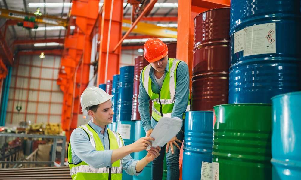 Two workers in hard hats and safety vests stand next to liquid storage drums. They both look at a paper one worker is holding.