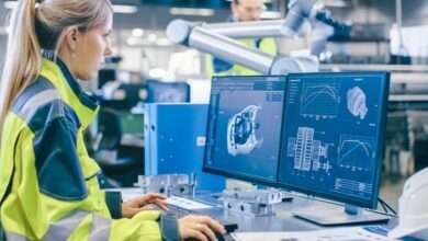 A woman wearing a yellow safety jacket stands behind a desk with two monitors. She is reviewing the digital specs for a part.