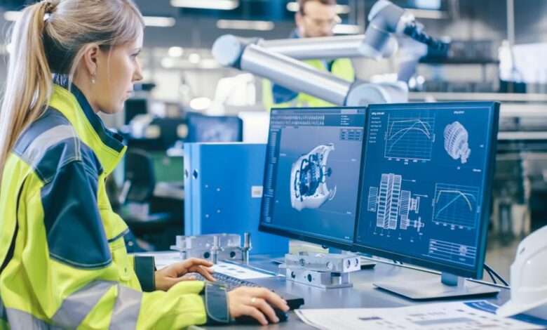 A woman wearing a yellow safety jacket stands behind a desk with two monitors. She is reviewing the digital specs for a part.