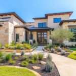 A modern home sits behind beautiful, well-maintained landscaping with trees, plants, stones, and hardscaping features.