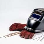 A welding helmet, a pair of well-worn red gloves, and several tungsten rods arranged against a clean white backdrop.