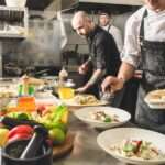 Three restaurant workers stand near a counter full of pasta dishes, kitchen equipment, and fresh produce.