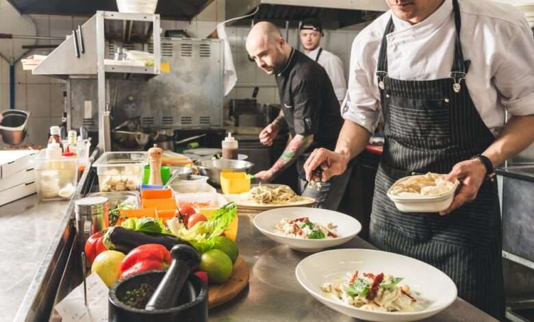 Three restaurant workers stand near a counter full of pasta dishes, kitchen equipment, and fresh produce.