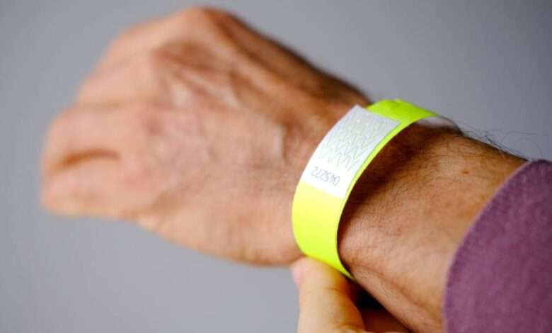 A close-up of a man's wrist with a purple shirt and a neon-yellow Tyvek paper wristband against a grey background.
