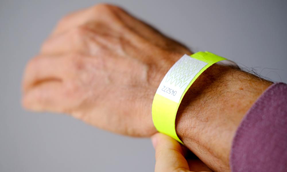 A close-up of a man's wrist with a purple shirt and a neon-yellow Tyvek paper wristband against a grey background.