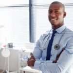 A doctor smiles as he stands in his office, wearing a shirt and tie. He has a stethoscope around his neck.