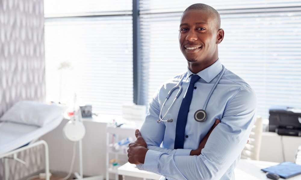 A doctor smiles as he stands in his office, wearing a shirt and tie. He has a stethoscope around his neck.