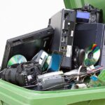 An open recycling bin with pieces of technology in it. There is a computer tower, a camera, and a laptop among the pile.