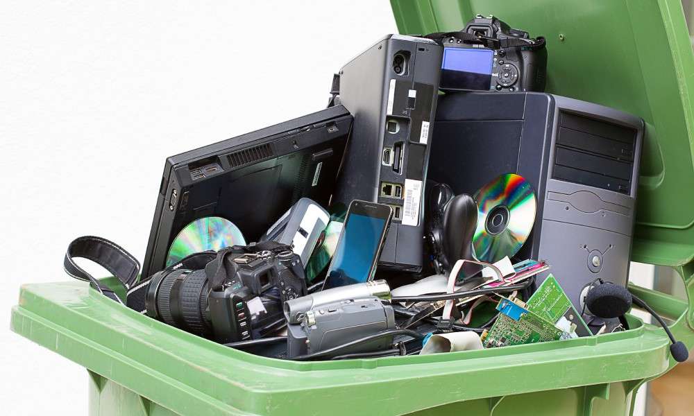 An open recycling bin with pieces of technology in it. There is a computer tower, a camera, and a laptop among the pile.