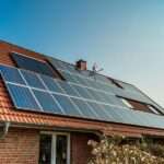 Numerous solar panels on the red roof of an older home. The exterior of the brick home is landscaped with bushes and plants.