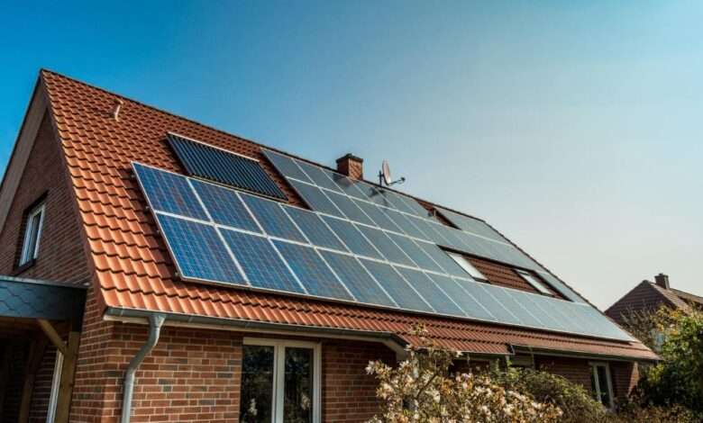 Numerous solar panels on the red roof of an older home. The exterior of the brick home is landscaped with bushes and plants.