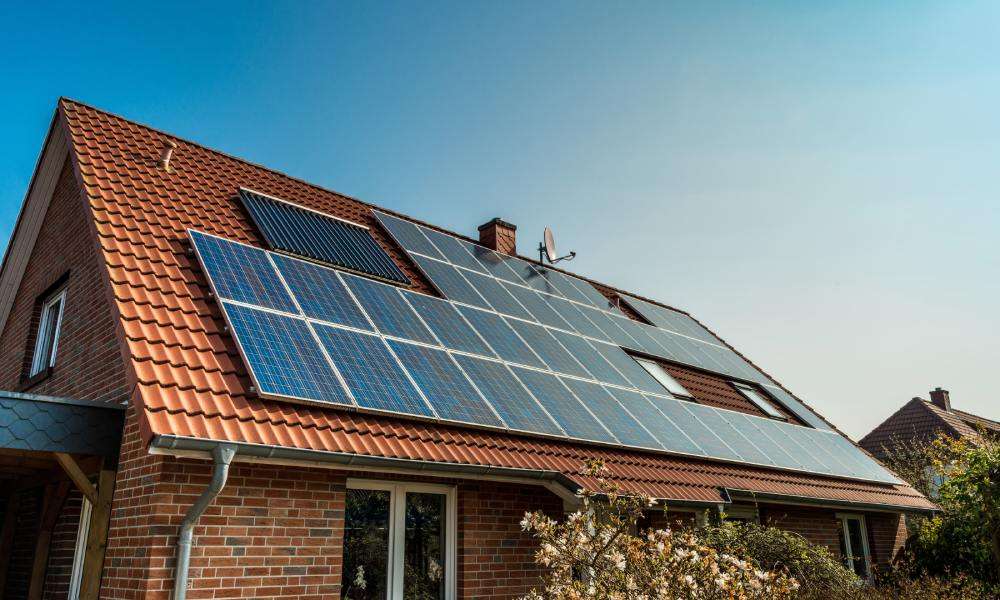 Numerous solar panels on the red roof of an older home. The exterior of the brick home is landscaped with bushes and plants.