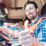 A trucker sitting in the cabin of his semi and giving a thumbs-up while smiling. He wears a blue flannel and an orange vest.