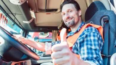 A trucker sitting in the cabin of his semi and giving a thumbs-up while smiling. He wears a blue flannel and an orange vest.