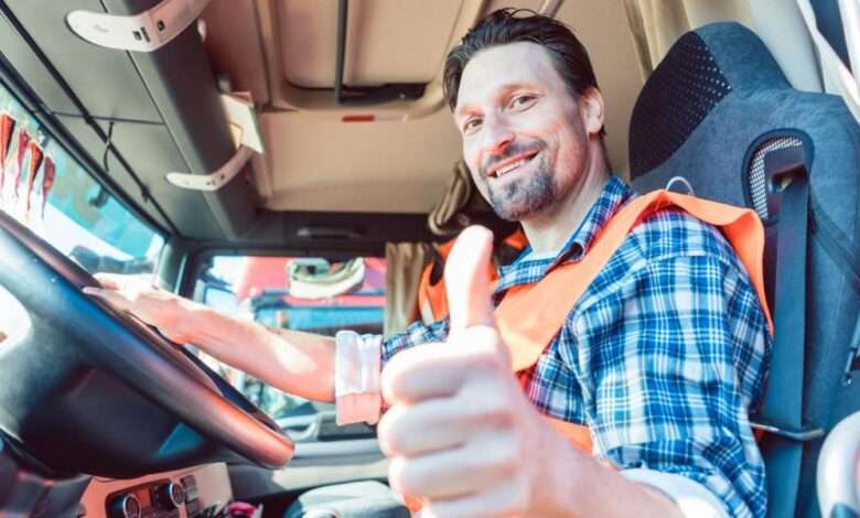 A trucker sitting in the cabin of his semi and giving a thumbs-up while smiling. He wears a blue flannel and an orange vest.