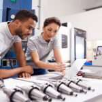 Two people, a man and a woman, are leaning over a desk and looking at a computer screen.