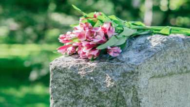Pink and purple flowers rest on a weathered gray headstone in a cemetery, with blurred green foliage in the background.