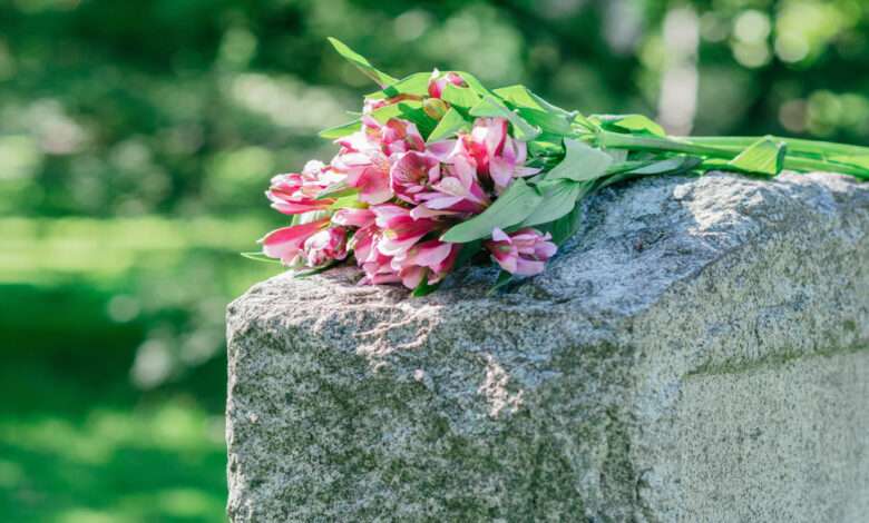 Pink and purple flowers rest on a weathered gray headstone in a cemetery, with blurred green foliage in the background.