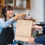 A woman with brown hair, wearing a denim shirt and black apron, hands a customer a brown paper bag at a restaurant.