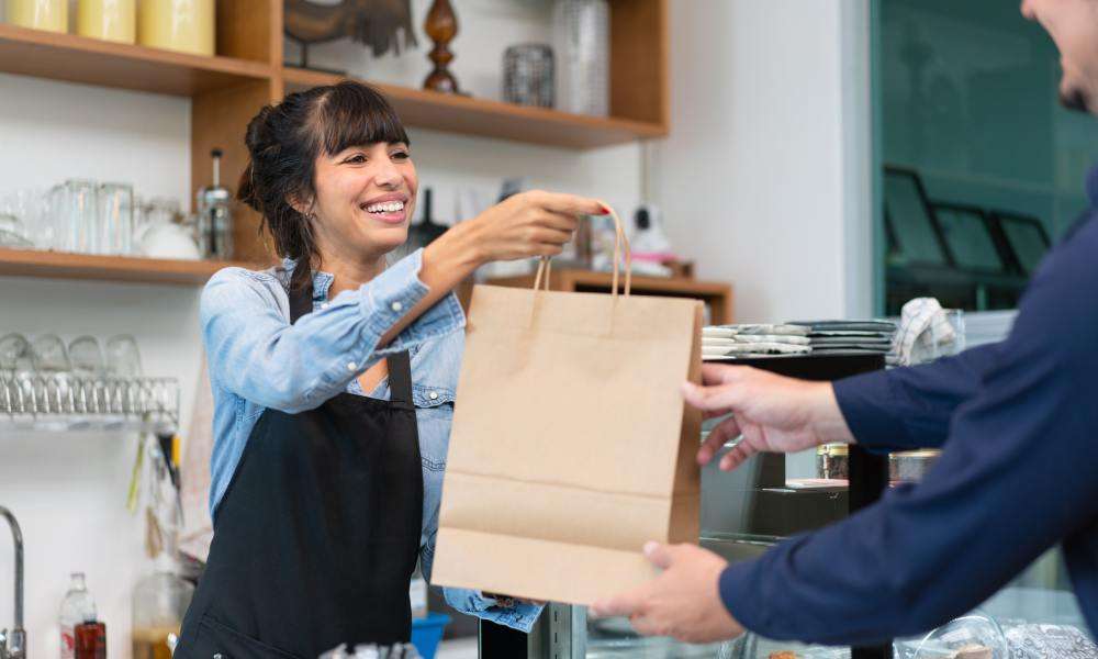 A woman with brown hair, wearing a denim shirt and black apron, hands a customer a brown paper bag at a restaurant.