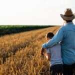 A farmer, wearing a cowboy hat, walks with his son through the vast fields of wheat on his farm. The sky is clear.