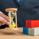 A close-up of a man holding a yellow-sand hourglass next to three miniature shipping containers stacked in a pyramid.