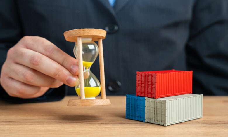 A close-up of a man holding a yellow-sand hourglass next to three miniature shipping containers stacked in a pyramid.