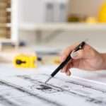 A close-up hand of an architect holding a pen, working on blueprint construction floor plans on a desk.