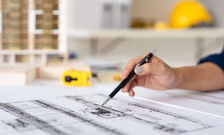 A close-up hand of an architect holding a pen, working on blueprint construction floor plans on a desk.