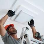 A person wearing a hard hat, gloves, and safety glasses stands on a ladder. They use a screwdriver to install a square light.