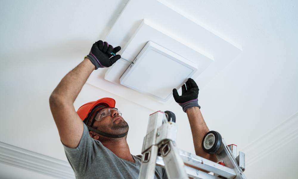 A person wearing a hard hat, gloves, and safety glasses stands on a ladder. They use a screwdriver to install a square light.
