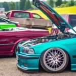 Several custom cars sit outside at an outdoor car exhibition. One blue-green vehicle has its hood open.