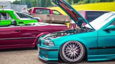 Several custom cars sit outside at an outdoor car exhibition. One blue-green vehicle has its hood open.