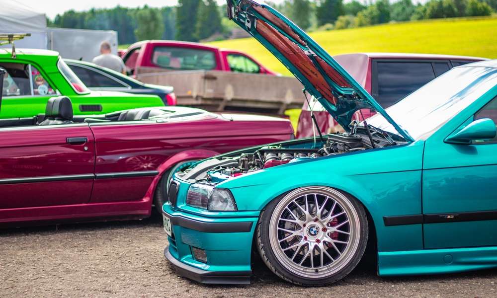 Several custom cars sit outside at an outdoor car exhibition. One blue-green vehicle has its hood open.