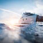 A large white and black catamaran motor yacht navigates the blue ocean as the sun sets in the background.