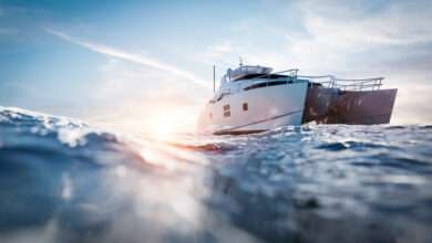 A large white and black catamaran motor yacht navigates the blue ocean as the sun sets in the background.