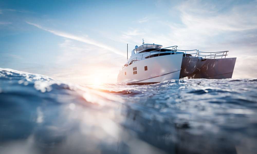 A large white and black catamaran motor yacht navigates the blue ocean as the sun sets in the background.