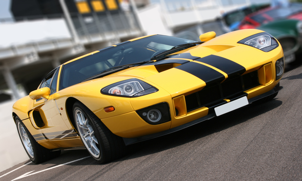 A yellow super car sits on a racing track, parked to other cars. Several buildings stand in the background.