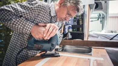 An older man in a plaid shirt uses a jigsaw to cut a wooden countertop inside a camper van with a built-in sink
