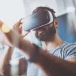 A bearded young man in a grey t-shirt reaches forward while wearing virtual reality goggles in a modern apartment.