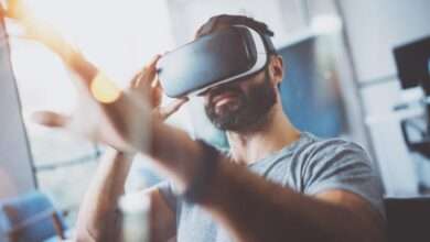 A bearded young man in a grey t-shirt reaches forward while wearing virtual reality goggles in a modern apartment.