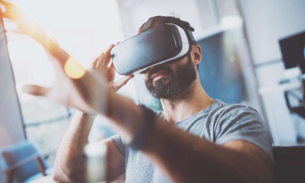 A bearded young man in a grey t-shirt reaches forward while wearing virtual reality goggles in a modern apartment.